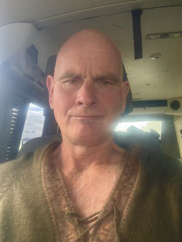 Ein glatzköpfiger Mann mit ernster Miene sitzt in einem Fahrzeug. Er trägt einen braunen Pullover. Durch ein Fenster dringt Sonnenlicht ein, das sein Gesicht leicht erhellt.