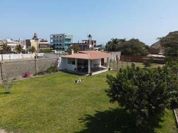 A small white house with a red-tiled roof sits in a spacious green yard, surrounded by a fence and trees, with urban buildings behind.