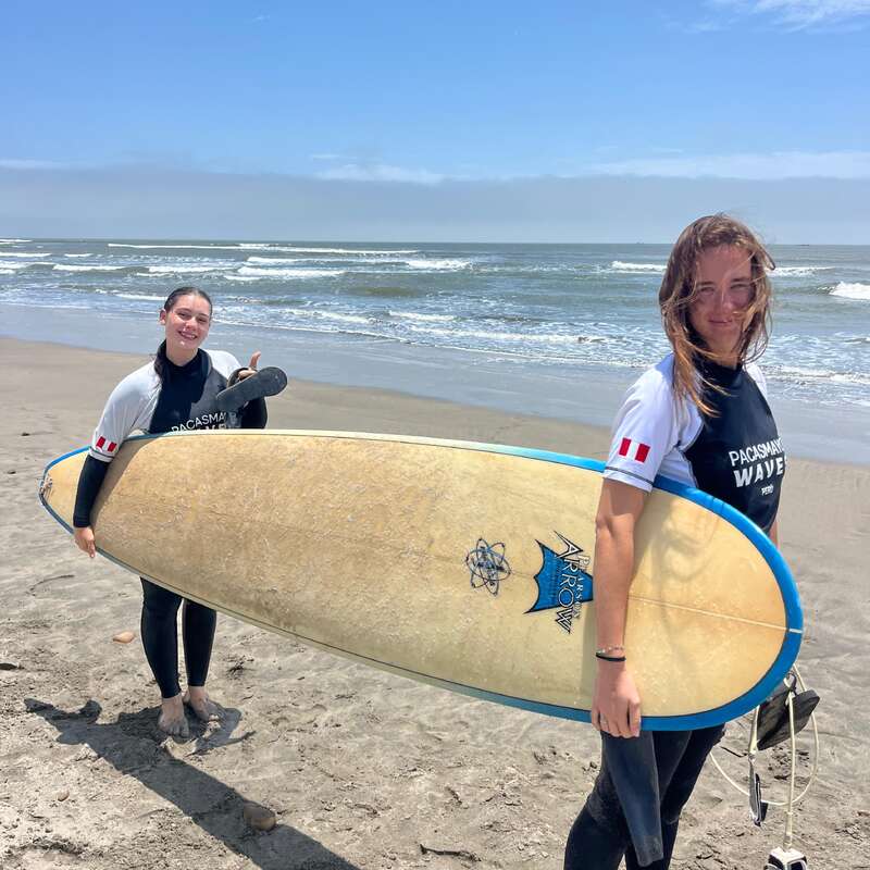 Two women in wetsuits, holding a surfboard, stand on a sunny beach near the ocean. Both are smiling, enjoying a day of surfing together.