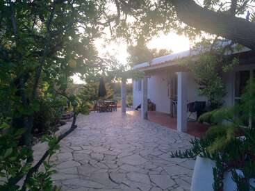 A charming patio with stone flooring, surrounded by lush greenery. White house with a red-tiled roof, outdoor furniture, and trees creating a peaceful atmosphere at sunset.