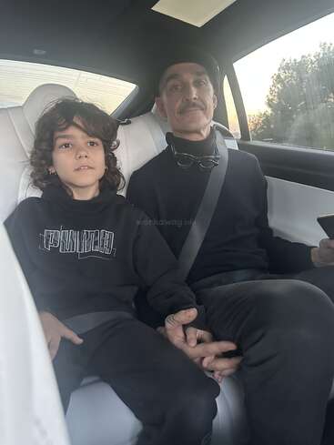 A young child and an adult sit together in the back seat of a car, holding hands, both wearing dark clothing and seat belts, looking calm.
