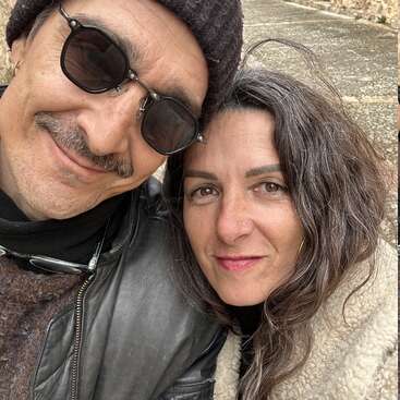 A smiling couple takes a close-up selfie outdoors. The man wears sunglasses, a hat, and a leather jacket. The woman has wavy hair and a nose ring.