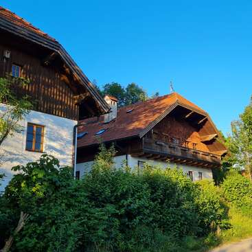 A charming rustic house with a red-tiled roof, wooden balcony, and white walls, surrounded by lush greenery under a bright blue sky. Peaceful scene.