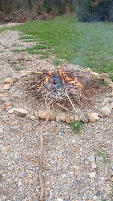 In einem Steinkreis auf felsigem Boden brennt ein kleines Lagerfeuer, umgeben von Gras und Wald, mit als Brennstoff aufgeschichteten Holzscheiten und aufsteigendem Rauch.