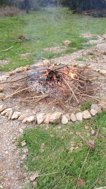 Ein kleines Feuer brennt auf einem Bett aus Zweigen, umgeben von einem Ring aus Steinen, draußen auf einem grasbewachsenen, teilweise felsigen Boden.