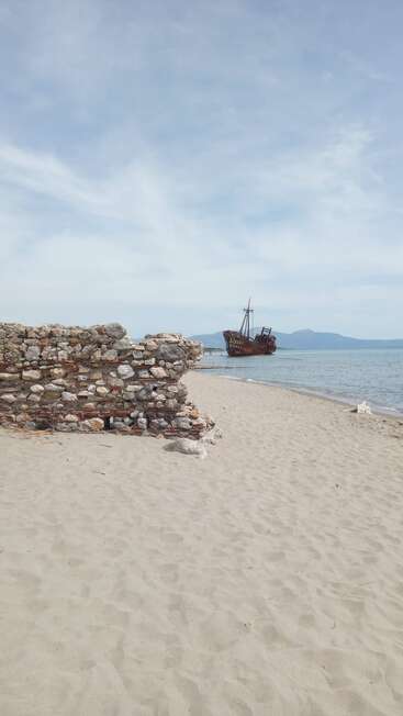 Ein Sandstrand mit alten Steinruinen im Vordergrund, ein rostiges, verlassenes Schiff in Ufernähe, ruhiges Meer und Berge, die im Hintergrund zu sehen sind.