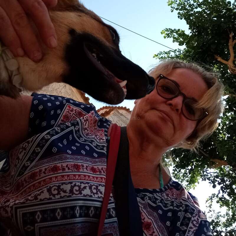 Eine Frau mit Brille und gemustertem Shirt macht draußen ein Selfie mit einem fröhlichen Hund. Hinter ihnen sind Bäume und ein sonniger, klarer Himmel zu sehen.