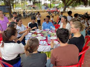 Eine Gruppe von Menschen sitzt um einen runden Tisch und genießt eine gemeinsame Mahlzeit im Freien. Im Hintergrund sind Teller, Wasserflaschen und einige Schaulustige zu sehen.