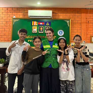 Five smiling young people pose inside a classroom. They make peace signs and playful gestures, standing in front of a green bulletin board with portraits and flags.