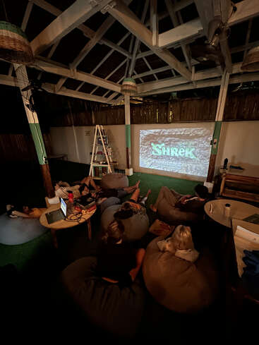Varias personas se relajan en pufs en una acogedora sala con techo abierto, viendo «Shrek» proyectada en la pared. La iluminación ambiental y los aperitivos crean un ambiente cómodo y acogedor.