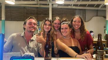 Cinco amigos posan felices juntos en un bar. Están sonrientes y relajados, con bebidas en la mano, lo que sugiere una velada divertida, sociable y alegre.