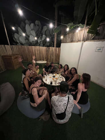 Un grupo de amigos se sienta alrededor de una mesa al aire libre por la noche, sonriendo y saludando con la mano, bajo luces de colores, disfrutando de unas bebidas y de la compañía mutua en un ambiente acogedor.