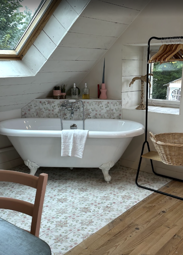 A cozy attic bathroom features a classic clawfoot bathtub, floral tiles, wooden ceiling, skylight, towel, plants, bottles, a hanging rack, and a woven basket.