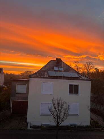 A white house stands under a dramatic, fiery orange sunset sky. Leafless trees are in front, highlighting the vibrant, colorful evening atmosphere in the neighborhood.