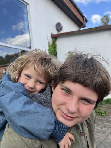 Two children pose outdoors; the younger one with curly blonde hair is playfully riding on the older one’s back. Both are smiling, enjoying a bright, sunny day.