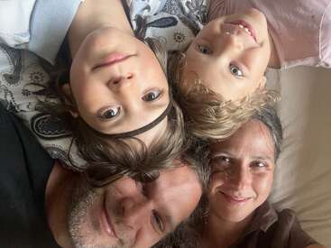 A joyful family of four lies close together, heads touching, smiling warmly at the camera. The mood is loving and relaxed, reflecting happiness and togetherness.