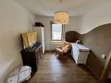 Dieses gemütliche Zimmer hat Holzböden, ein Fenster in der Mitte, Vintage-Möbel, einen Schaukelstuhl, eine Kommode und eine gemusterte braune Wand mit einer Pendelleuchte.