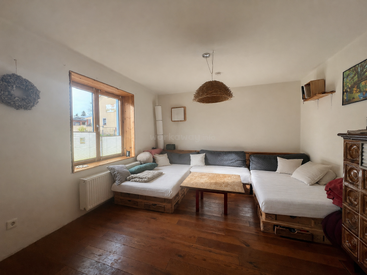 Dieses gemütliche Wohnzimmer besticht durch Palettensofas mit weißen Kissen, einen Holzboden, einen Tisch in der Mitte, ein großes Fenster, eine schlichte Einrichtung und eine geflochtene Hängelampe.