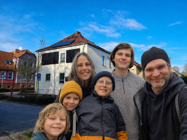 Une joyeuse famille de six personnes, emmitouflée dans des vêtements chauds et coiffée de bonnets, sourit ensemble à l'extérieur par une journée ensoleillée, avec en arrière-plan des maisons colorées et un ciel bleu limpide.
