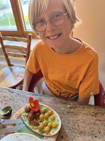 Uma criança sorridente, de óculos e camiseta laranja, está sentada à mesa, exibindo com orgulho um pão achatado colorido coberto de vegetais, decorado com tomates e uma flor comestível.