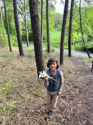 Um menino está em uma área arborizada, sorrindo e segurando um aviãozinho de brinquedo feito de papel e palitos. Ele está cercado por árvores e vegetação em uma encosta.
