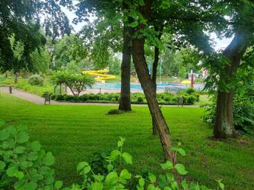 Um parque verdejante com árvores altas e gramado bem cuidado. Ao fundo, dá pra ver uma piscina com toboáguas amarelas brilhantes. Um ambiente tranquilo.