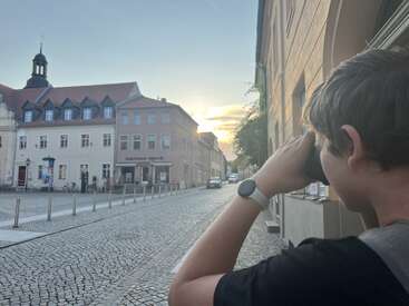 Um garoto de cabelo curto e com um relógio olha através de binóculos numa rua tranquila de paralelepípedos, observando prédios antigos em estilo europeu durante um pôr do sol pitoresco.