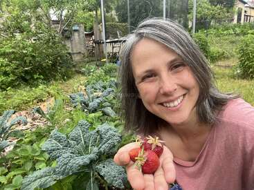 Eine lächelnde Frau mit grauen Haaren hält frisch gepflückte Erdbeeren in der Hand, während sie in einem üppigen Garten voller grüner Blattgemüse und Pflanzen posiert.