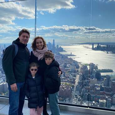 The image depicts a family of four posing for a photo on an observation deck, with a stunning cityscape and river view behind them.