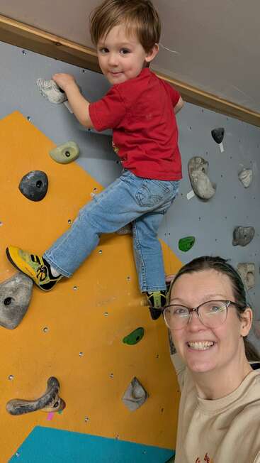 Uma criança sorridente com uma camisa vermelha escala uma parede de escalada interna, enquanto um adulto de óculos sorri orgulhosamente ao lado dela, capturando um momento alegre e aventureiro juntos.