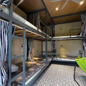 Cette image montre une chambre d'auberge de jeunesse moderne avec quatre lits superposés, des rideaux d'intimité, un sol carrelé à motifs, des lampes de lecture, un plafond en bois et une chaise hamac vert vif.