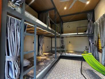 This image shows a modern hostel dormitory room with four bunk beds, privacy curtains, patterned tile floor, reading lights, wooden ceiling, and a bright green hammock chair.