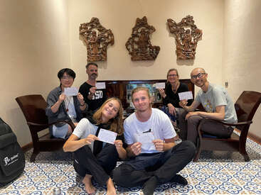 Six people sit together in a warmly lit room, holding cards and smiling. They appear relaxed and happy, surrounded by ornate wall art and patterned tiles.