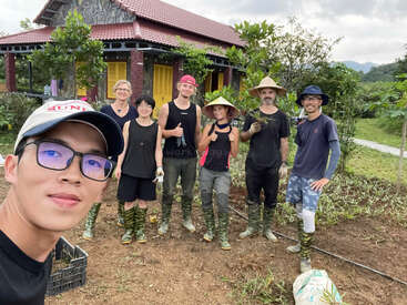 A group of six people pose for a photo outdoors, all wearing boots and casual clothes, standing near a house with plants and trees around them.