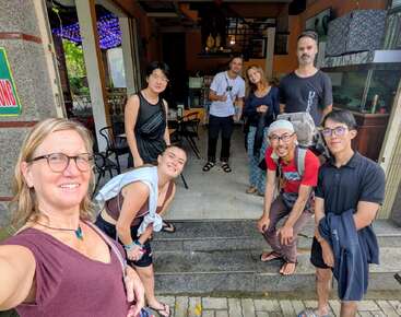 A group of eight people pose happily in front of a café or restaurant, with some smiling and bending forward. The atmosphere appears friendly and welcoming.