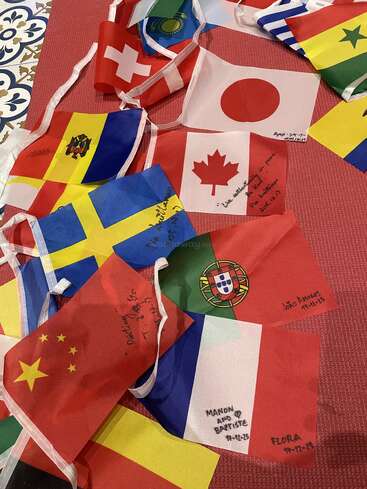 A collection of various country flags is spread out on a surface. Some flags have handwritten names or messages, creating a colorful, international, and personalized display.