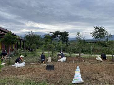A group of people are weeding or planting in a garden beside a house. Lush green scenery and distant mountains are visible under a cloudy sky.