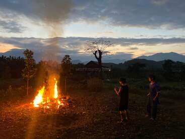 Two people stand near a bright bonfire at dusk, surrounded by nature and mountains under a dramatic cloudy sky, creating a peaceful, atmospheric outdoor scene.