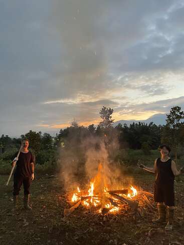 Two people in black clothes stand near a large campfire at dusk, surrounded by trees and a cloudy sky, creating a warm, rustic, and dramatic atmosphere.