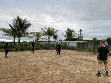 Six people stand on a sandy volleyball court surrounded by palm trees, under a cloudy sky, appearing ready to play. Tropical, relaxed outdoor scene, casual attire.