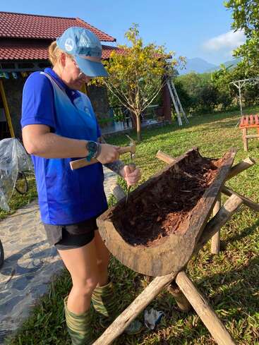 Una persona vestida con una camisa azul, una gorra y botas talla un gran tronco de madera al aire libre, utilizando herramientas, rodeada de vegetación, con una casa y unas montañas al fondo.