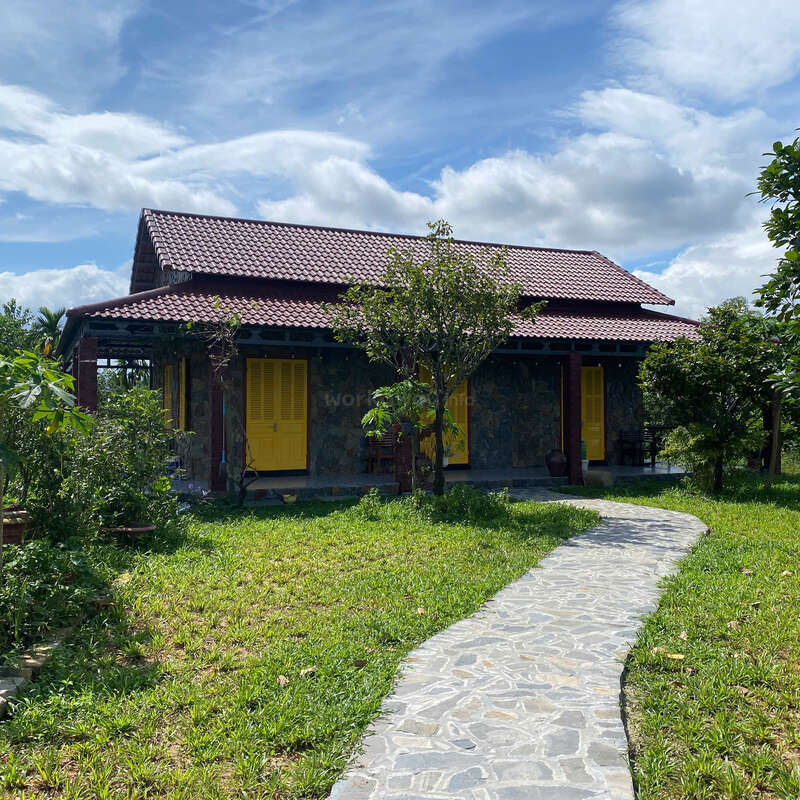 Une charmante maison rustique au toit de tuiles rouges et aux portes jaune vif se trouve au milieu d'une verdure luxuriante. Un sentier en pierre mène à travers le jardin animé.