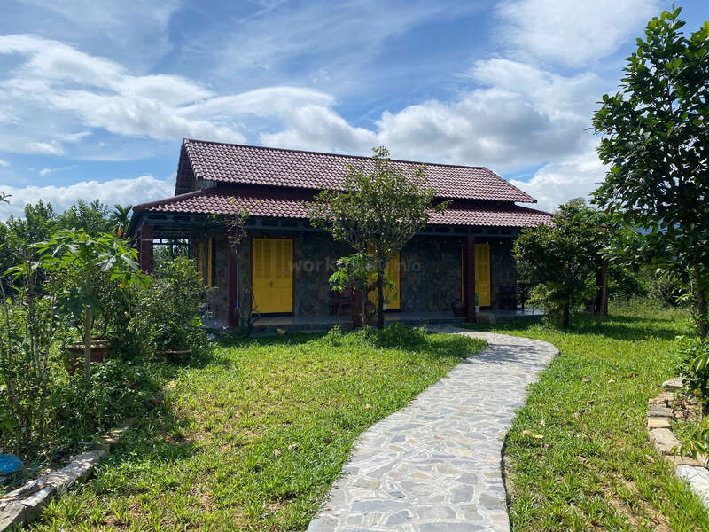 A charming, rustic house with a red-tiled roof and bright yellow doors sits amid lush greenery, a stone pathway leads through the vibrant garden.