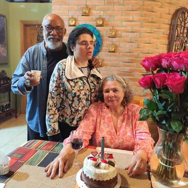 Três pessoas comemoram juntas em um ambiente fechado. Uma mulher está sentada diante de um bolo de aniversário e flores, segurando vinho; duas pessoas estão atrás dela, sorrindo, criando uma atmosfera alegre e amorosa.