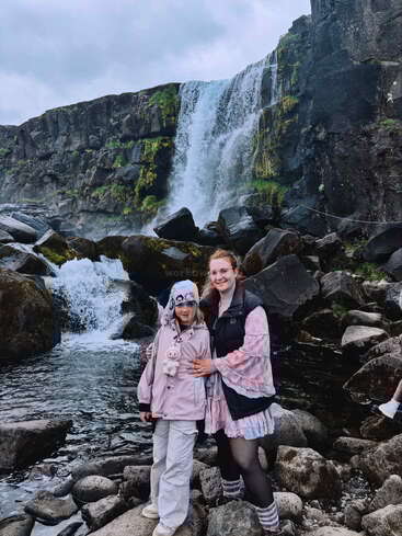 Une femme et une fille posent ensemble sur un terrain rocailleux devant une belle cascade, toutes deux habillées chaudement et souriantes, profitant d'une aventure dans la nature.