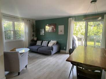 Dieses gemütliche Wohn-Esszimmer hat eine grüne Akzentwand, ein graues Sofa, Kunstwerke, einen Esstisch aus Holz, große Fenster, Terrassentüren und viel natürliches Sonnenlicht.