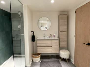 In diesem modernen Badezimmer sorgen eine Glasdusche, ein runder Spiegel, ein Waschtisch und ein Schrank aus hellem Holz, ein Plüschhocker, ein geflochtener Korb und neutrale Töne für eine klare, einladende Atmosphäre.