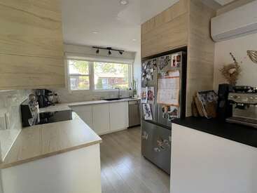 Moderne Küche mit hellen Holzschränken, Geräten aus Edelstahl und großem Fenster. Im Kühlschrank hängen Familienfotos. Die Kaffeemaschine, die minimalistische Einrichtung und das natürliche Licht schaffen eine gemütliche Atmosphäre.