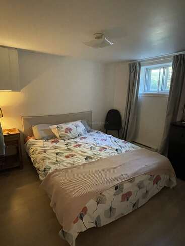 Ein gemütliches Schlafzimmer mit einem Doppelbett mit geblümter Bettwäsche, einem Nachttisch mit einer Lampe, einem Fenster mit Vorhängen, einem Stuhl und minimalistischen Möbeln auf Hartholzböden.