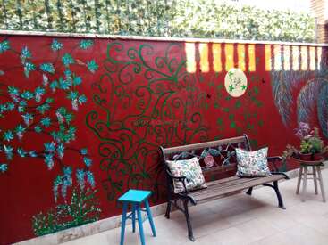 Un colorido patio presenta una pared roja pintada con enredaderas y flores verdes, un banco de madera con cojines florales, un taburete azul y plantas en macetas.